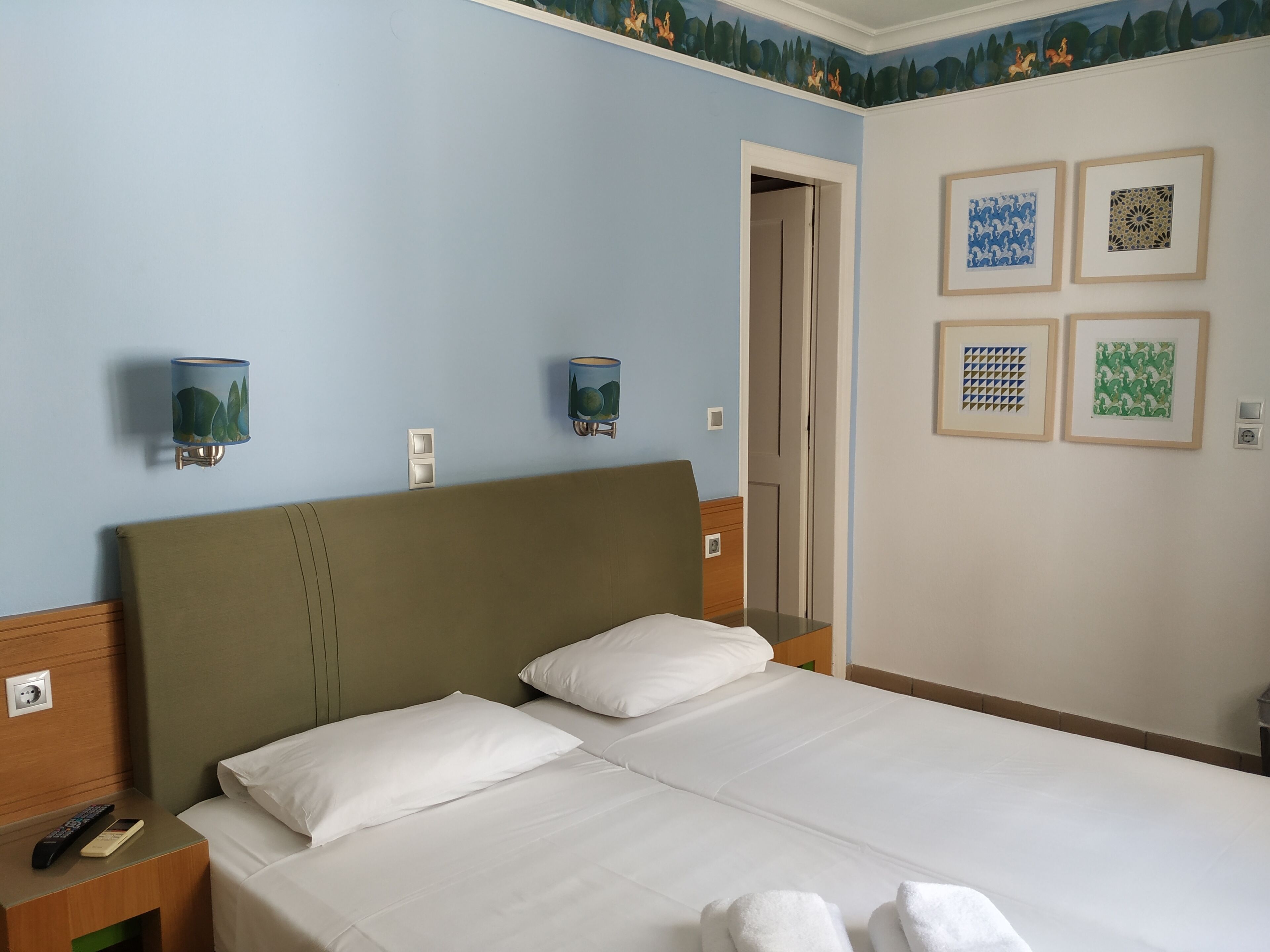 A hotel room with a bed, two wall-mounted lamps, a door, and four framed artworks on the wall.