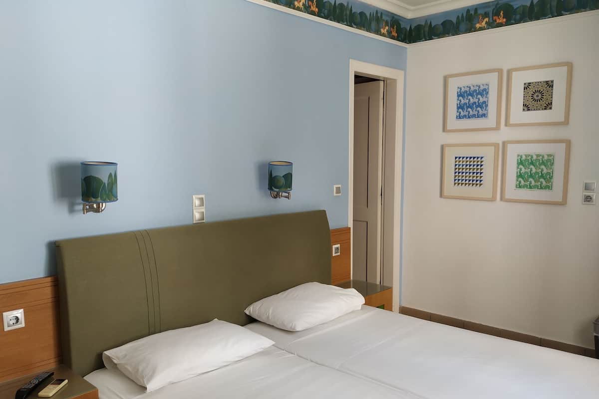 A hotel room with a bed, two wall-mounted lamps, a door, and four framed artworks on the wall.