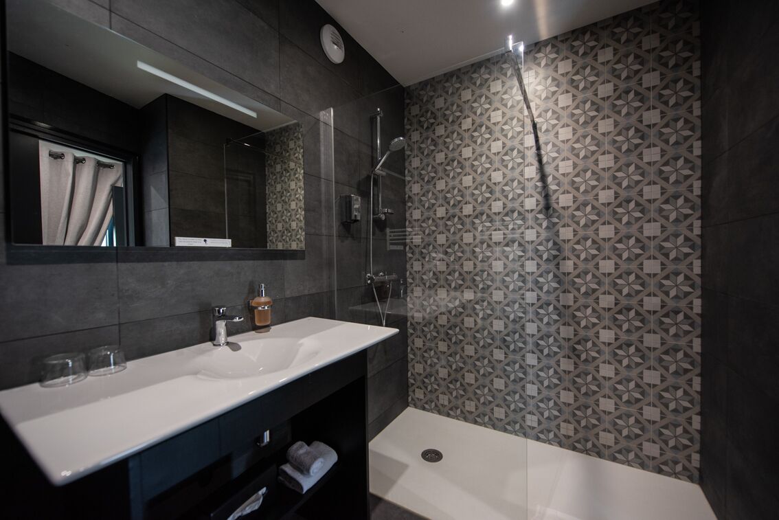 Une salle de bain moderne avec un lavabo blanc, des carreaux foncés, une douche et un mur orné d’un motif.