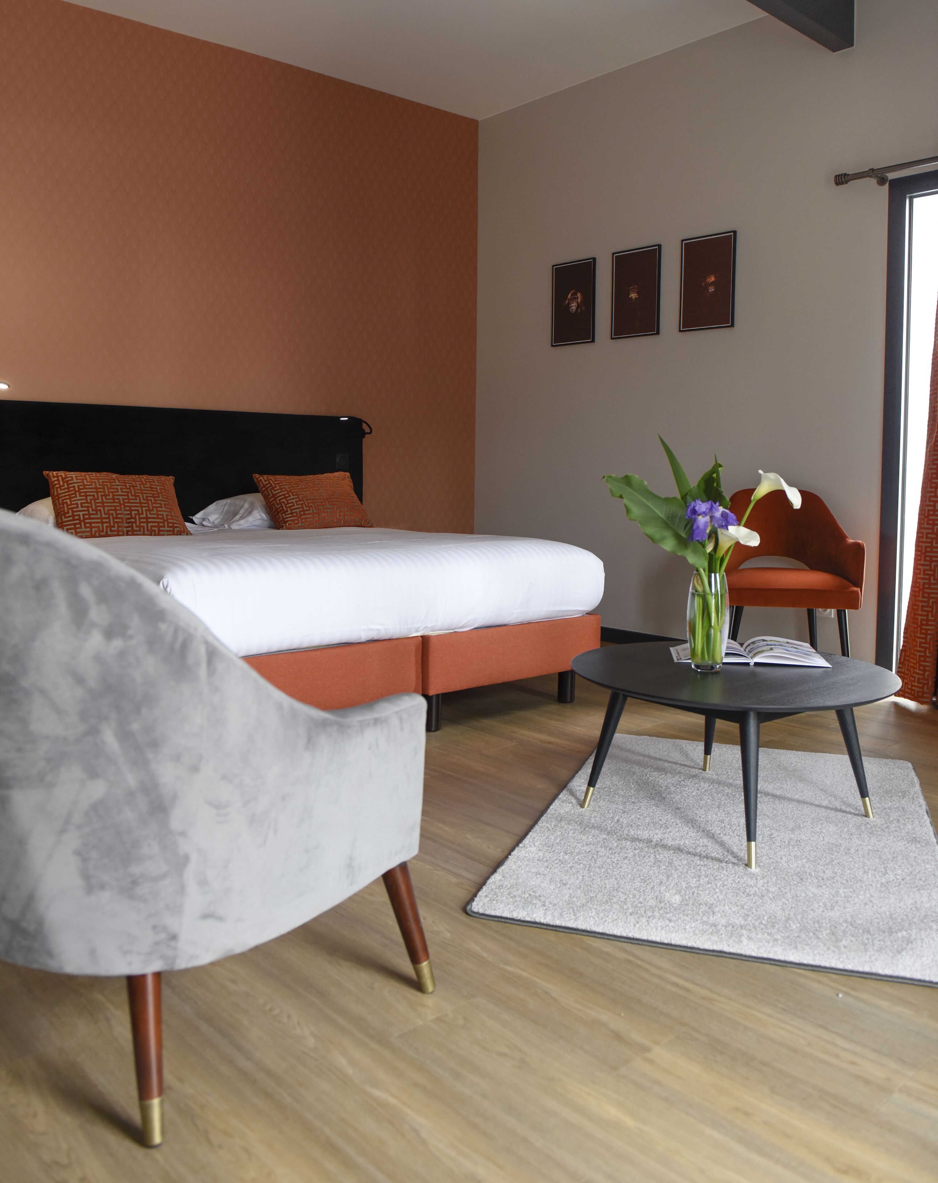 Una habitación de hotel moderna con una cama, un sillón gris, una mesita redonda con un florero y un sofá rojo.