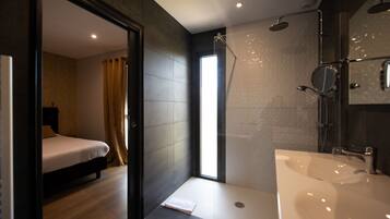 Un baño moderno con un lavamanos grande, ducha y una cama visible a través de un arco.