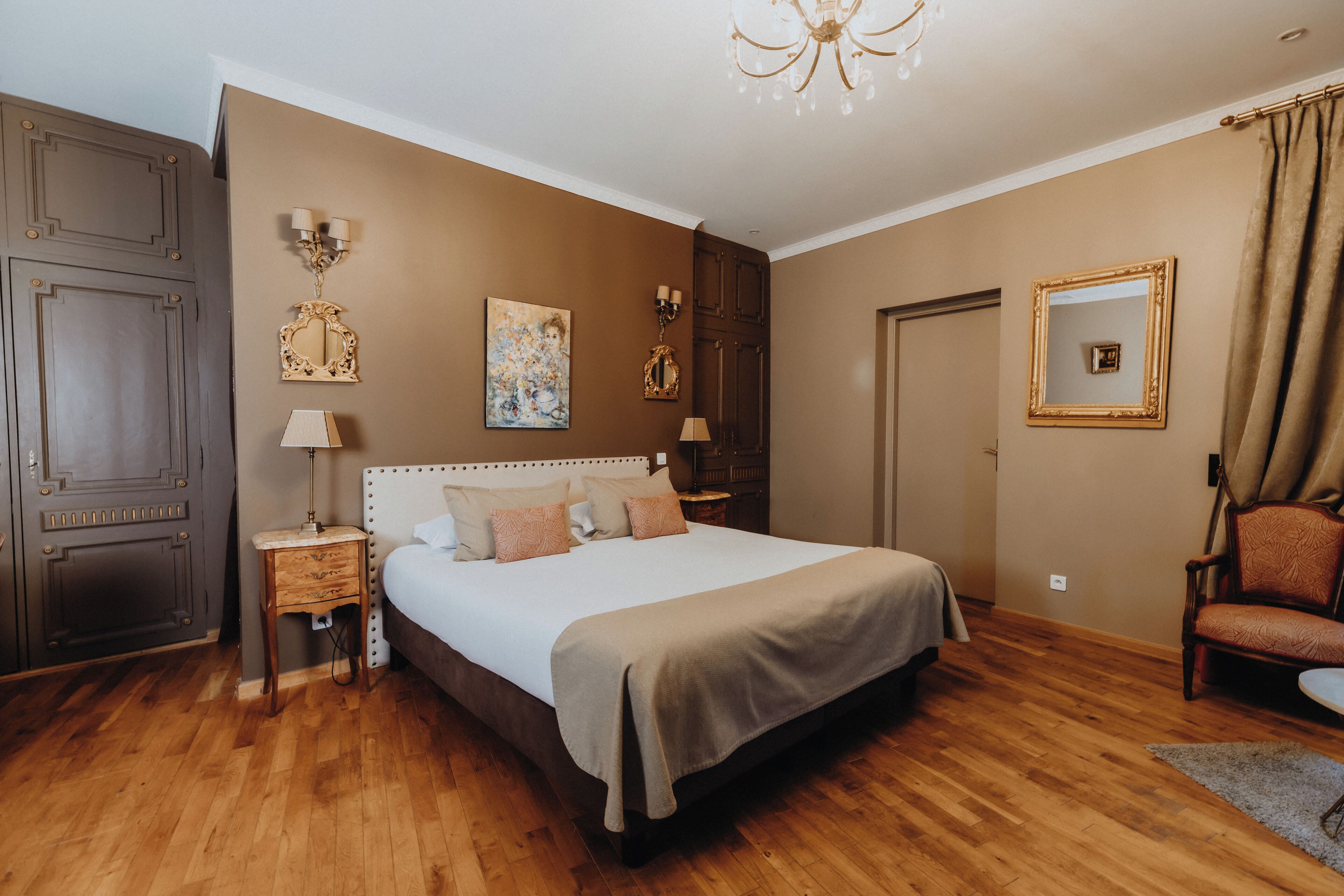 A bedroom with a large bed, wooden floor, dark wardrobe, bedside tables, and a chandelier.