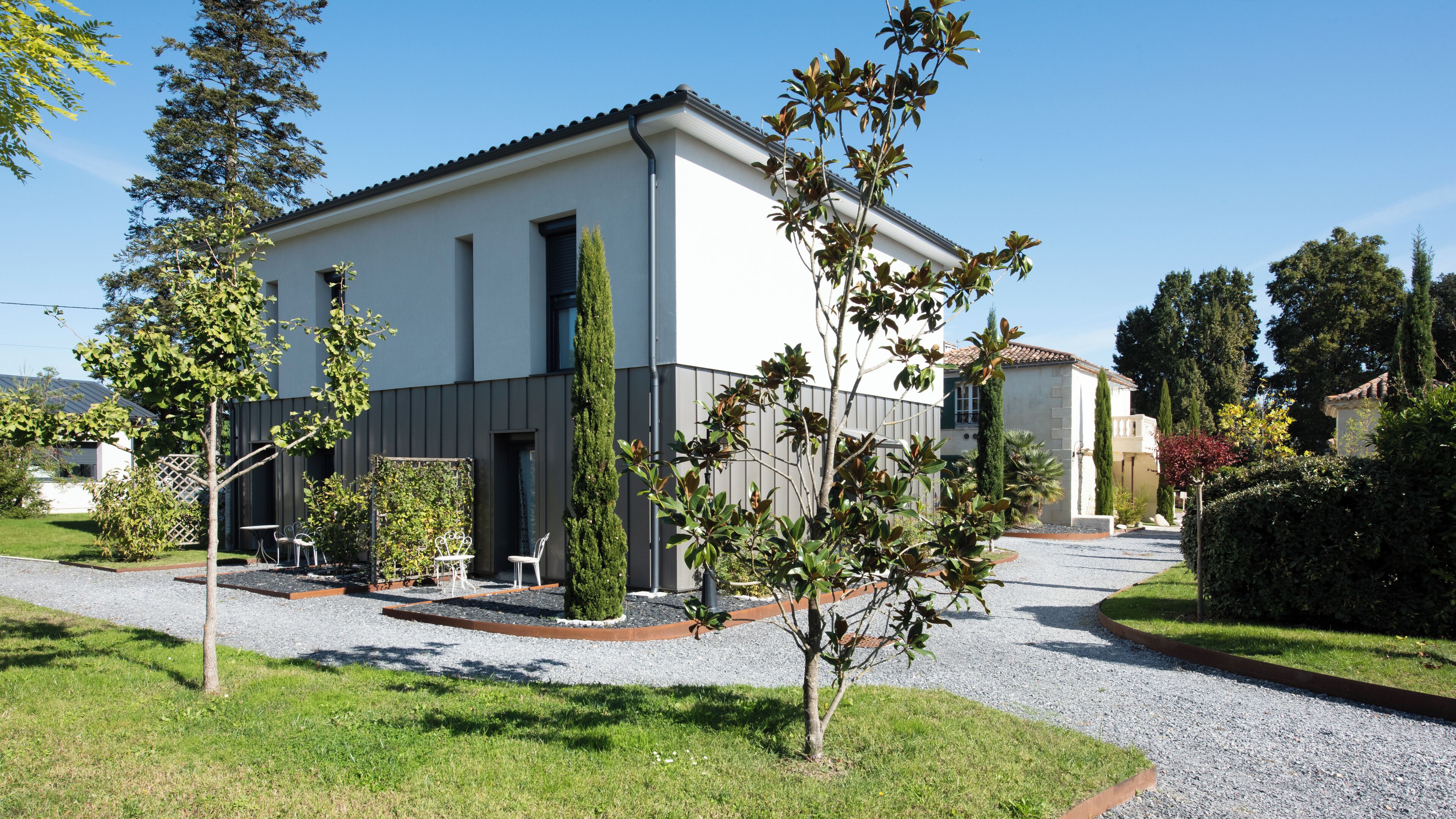 A modern two-story building with a white facade, a gravel driveway, and a small garden area with trees and shrubs.