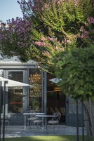 Un área de estar al aire libre con mesas y sillas, un acceso por puerta de vidrio y una planta ornamental con flores rosas.