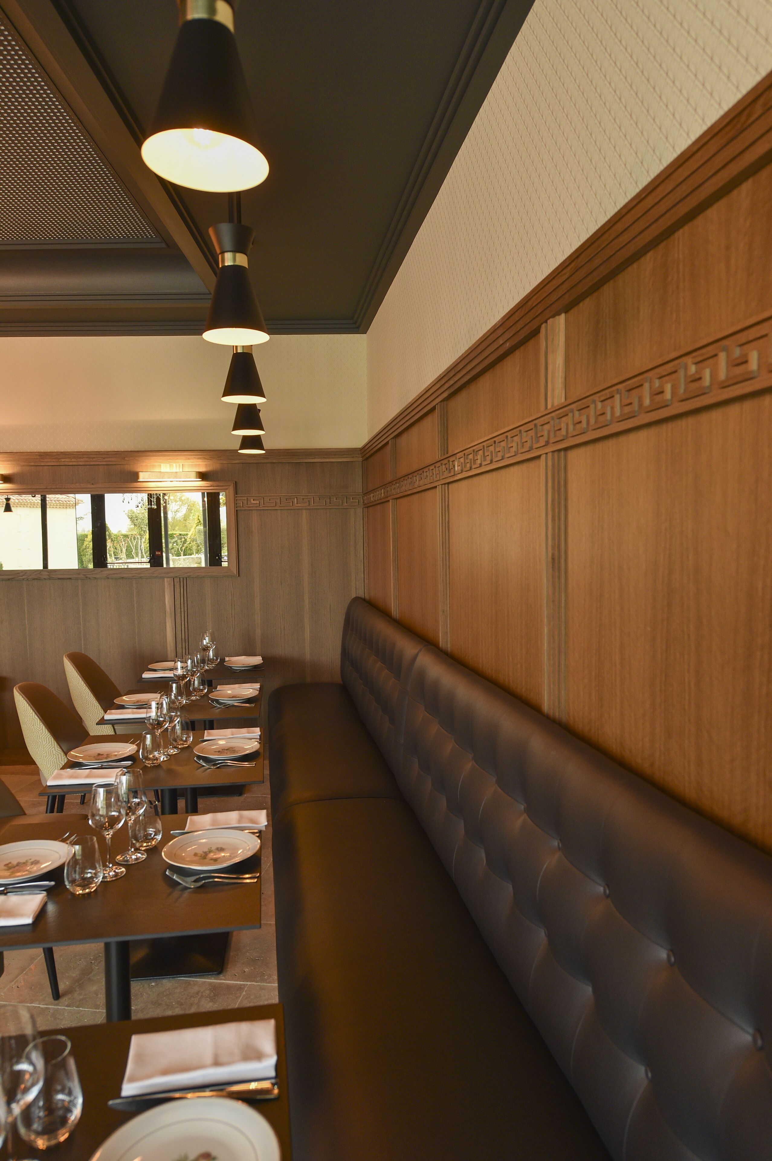 A long dining table set with plates, glasses, and cutlery, flanked by cushioned seating and pendant lighting.