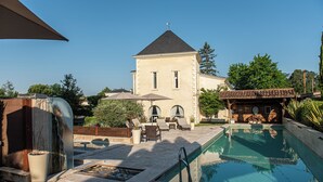 A pool area with a lounge chair and umbrella, a stone building with a clock, and a wooden structure with a clock. - Domaine de Larchey & son restaurant - Bordeaux Sud (Saint-Medard-d'Eyrans)