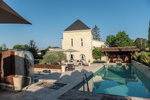 Un área de piscina con una silla de descanso y una sombrilla, un edificio de piedra con un reloj, y una estructura de madera también con un reloj.