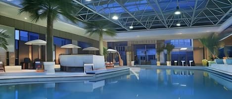An indoor swimming pool with lounge chairs, palm trees, and a high ceiling with a skylight.