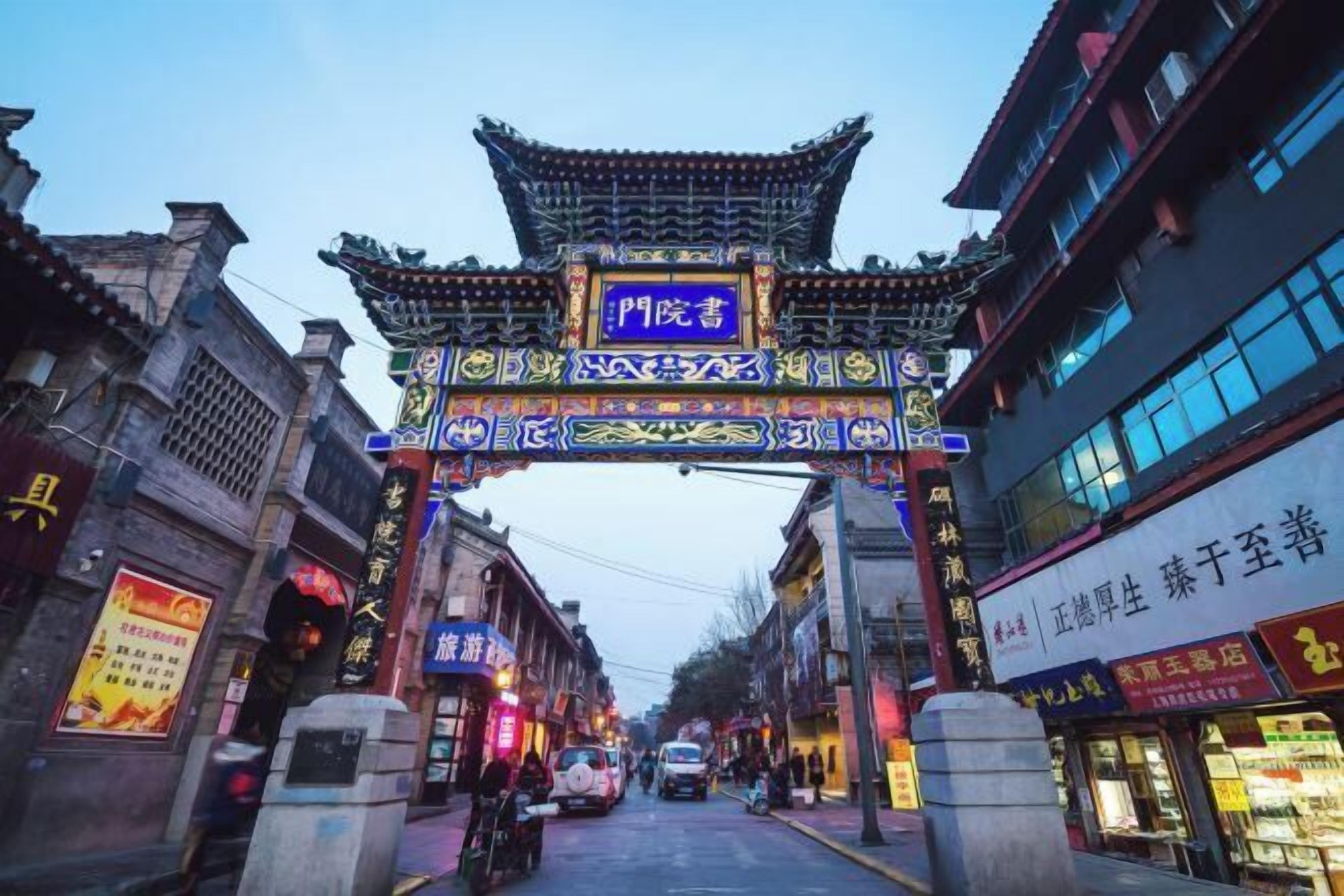 Um portão chinês tradicional com desenhos e caracteres elaborados, dando acesso a uma rua repleta de lojas e edifícios.