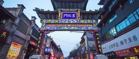 Um portão chinês tradicional com desenhos e caracteres elaborados, dando acesso a uma rua repleta de lojas e edifícios.