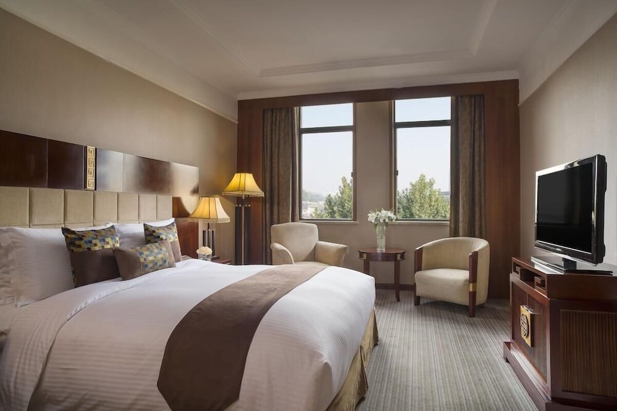 Ein Hotelzimmer mit einem großen Bett, einem Fernseher, einem Schreibtisch mit Lampe, zwei Stühlen und Blick ins Freie durch die Fenster.