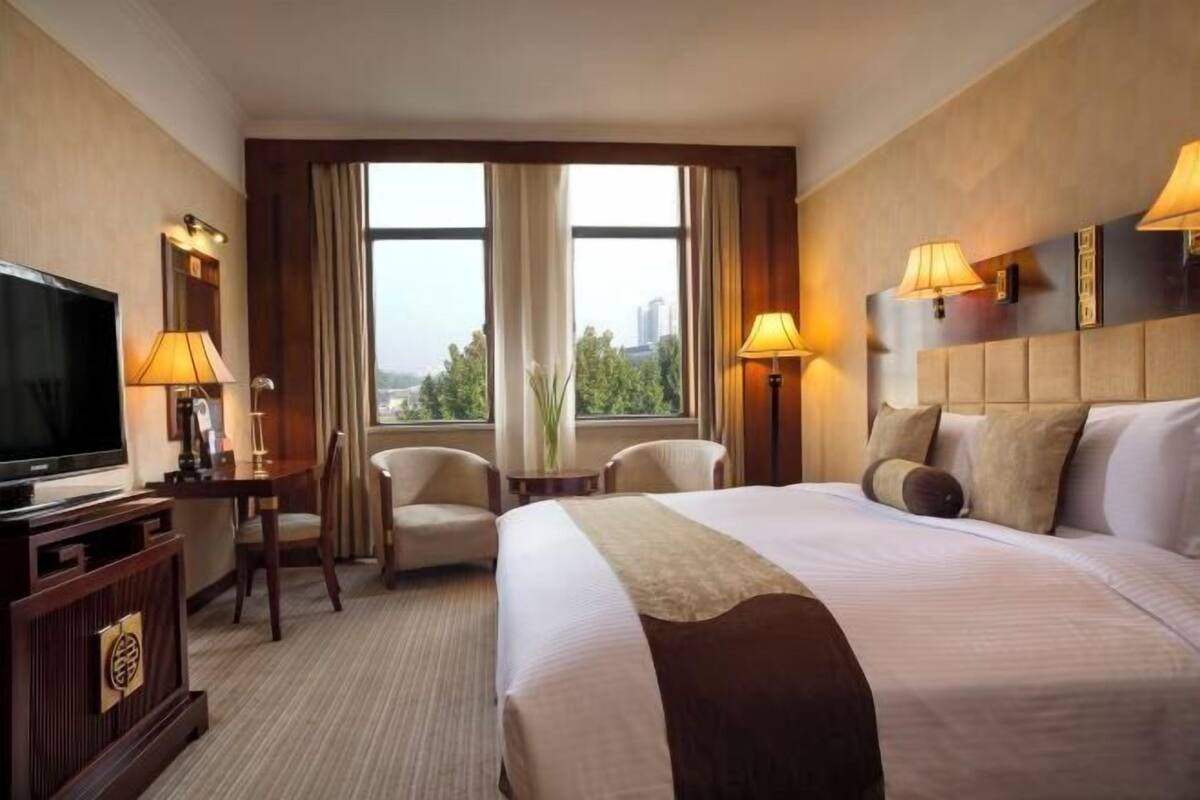 Habitación de hotel con una cama grande, televisor, dos sillas, una mesita y vistas a la ciudad a través de amplias ventanas.