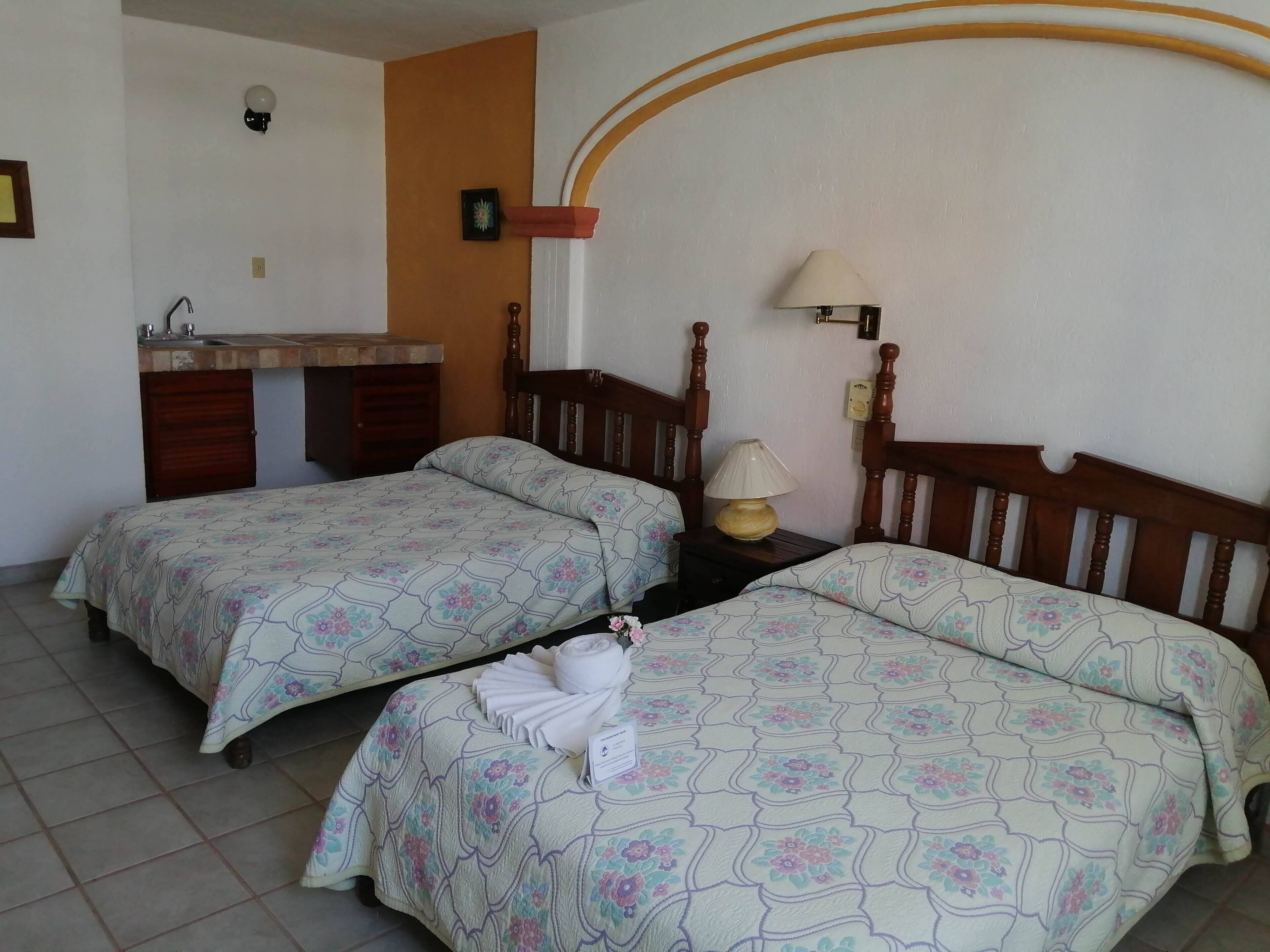 A room with two beds, a small bedside table with a lamp, a wall-mounted light, and a bathroom with a sink and mirror.