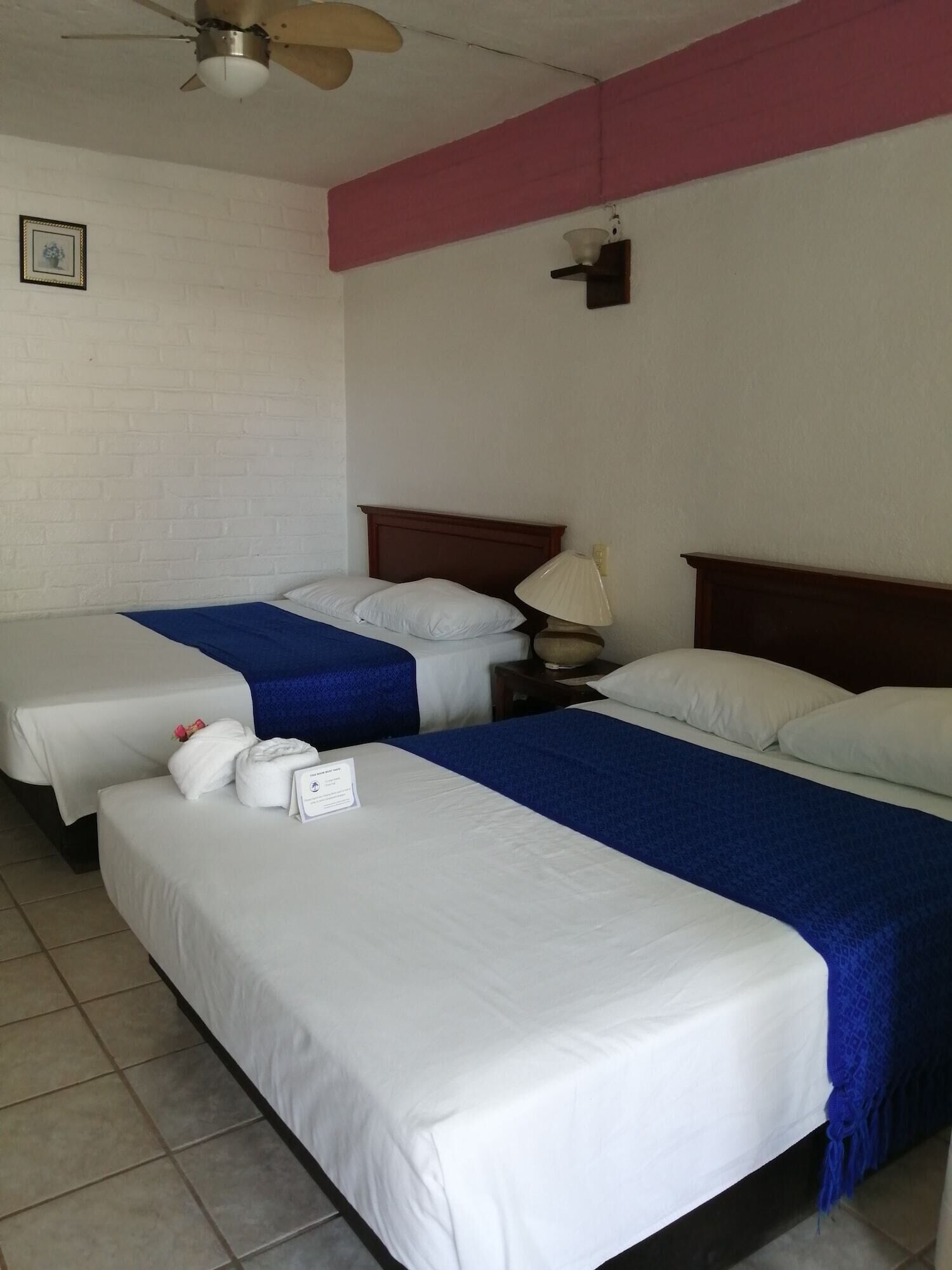 Quarto de hotel com duas camas, cada uma com roupa de cama branca e colchas azuis. Há um ventilador de teto, uma luminária na parede e um quadro emoldurado.
