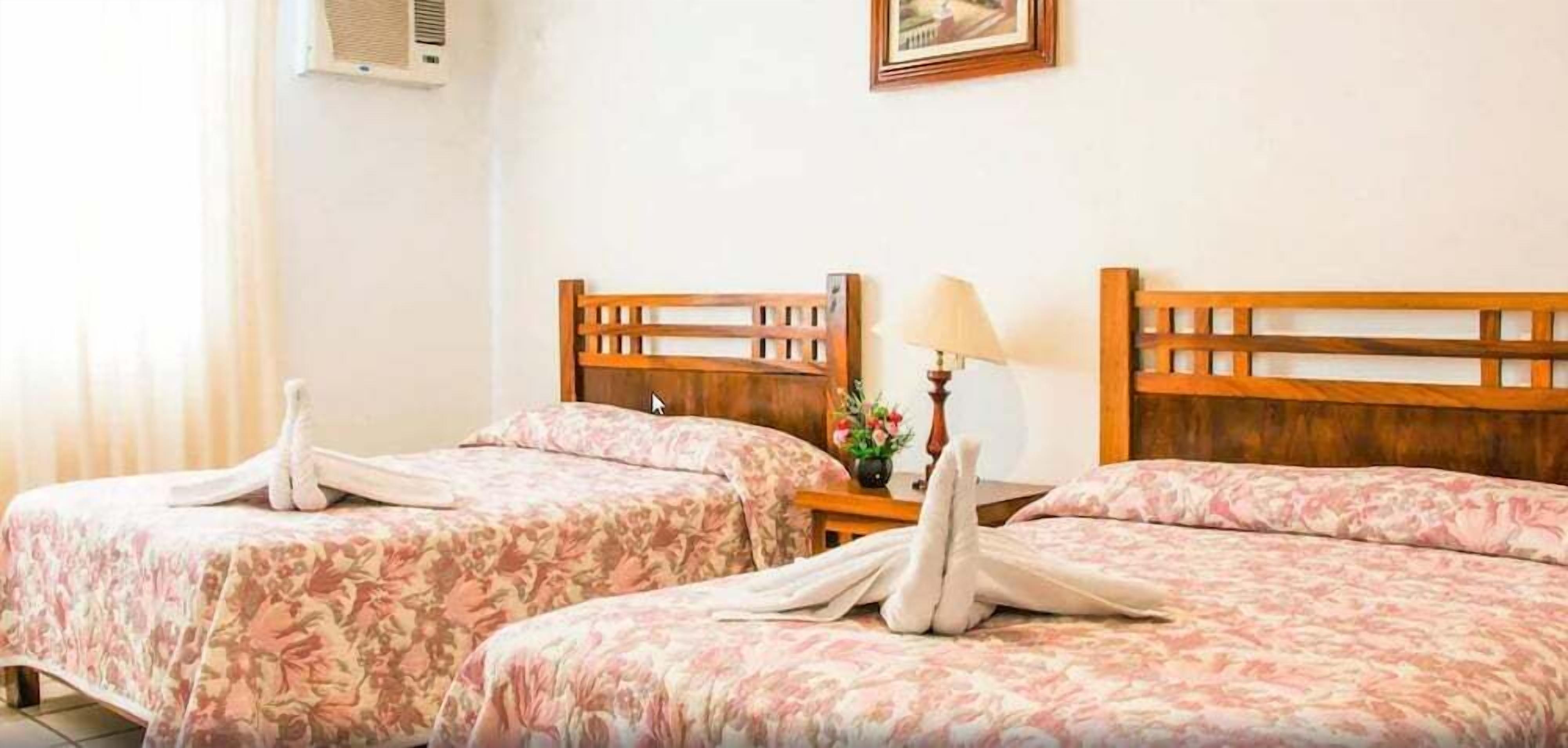 A hotel room with two beds, each with a wooden headboard and floral bedspread. There is a bedside table with a lamp and a flower vase.