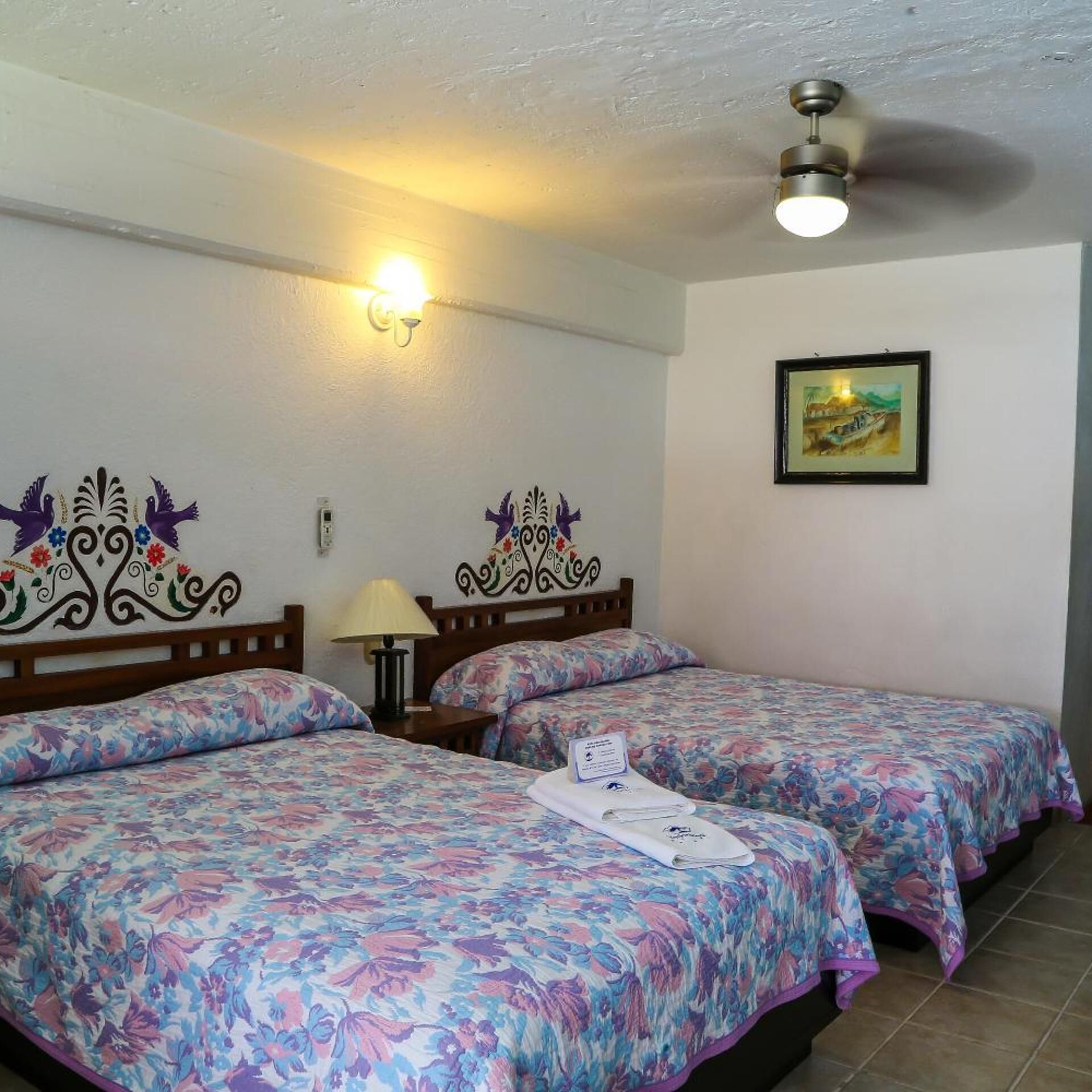 A hotel room with two beds, a ceiling fan, a wall-mounted light, and a framed painting.
