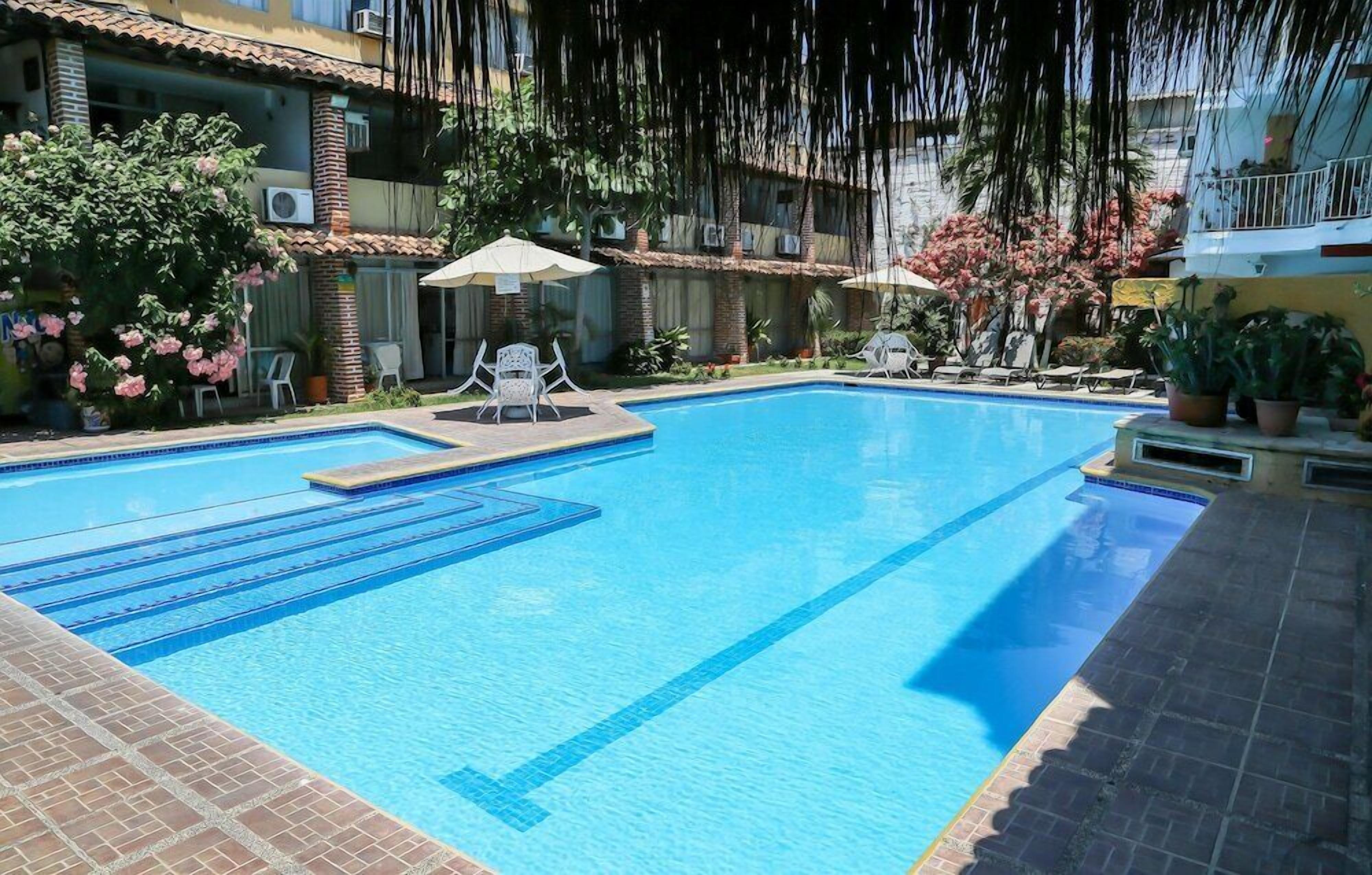 A swimming pool with lounge chairs and umbrellas, surrounded by buildings with balconies and potted plants.