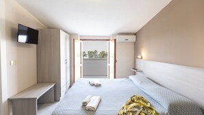 A modern hotel room with a bed, a TV mounted on the wall, a bench, and an open door leading to a balcony with a view of trees.