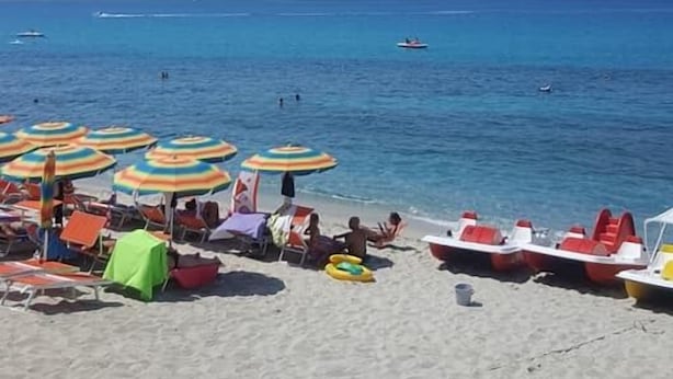 A beach with rows of colorful umbrellas and lounge chairs, a few people, and a boat in the water.