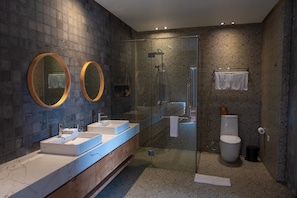 A modern bathroom with a glass-enclosed shower, two sinks with marble countertops, and two round mirrors. - Tegal Sari Accommodation (Ubud)