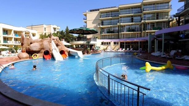 A swimming pool with slides and a rock formation, surrounded by lounge chairs and a building with balconies.