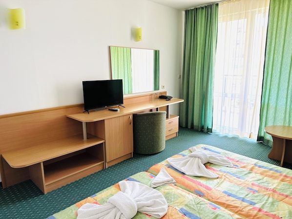 Ett hotellrum med ett skrivbord i trä, en tv, en spegel, en grön gardin, en säng med handdukar och en grön matta.