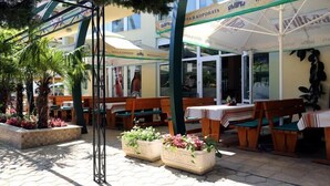 An outdoor cafe with wooden tables and benches, surrounded by potted plants and umbrellas.