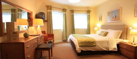 A hotel room with a bed, a desk with a chair, a dresser, a mirror, a bedside table with a lamp, and a window with curtains.
