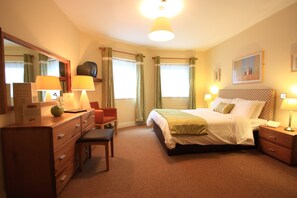 A hotel room with a bed, a desk with a chair, a dresser, a mirror, a bedside table with a lamp, and a window with curtains. - O'donnabhain's Guesthouse (Kenmare)