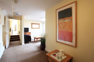 Habitación de hotel con pasillo alfombrado, un escritorio con cafetera, una silla, una planta y un cuadro en la pared.