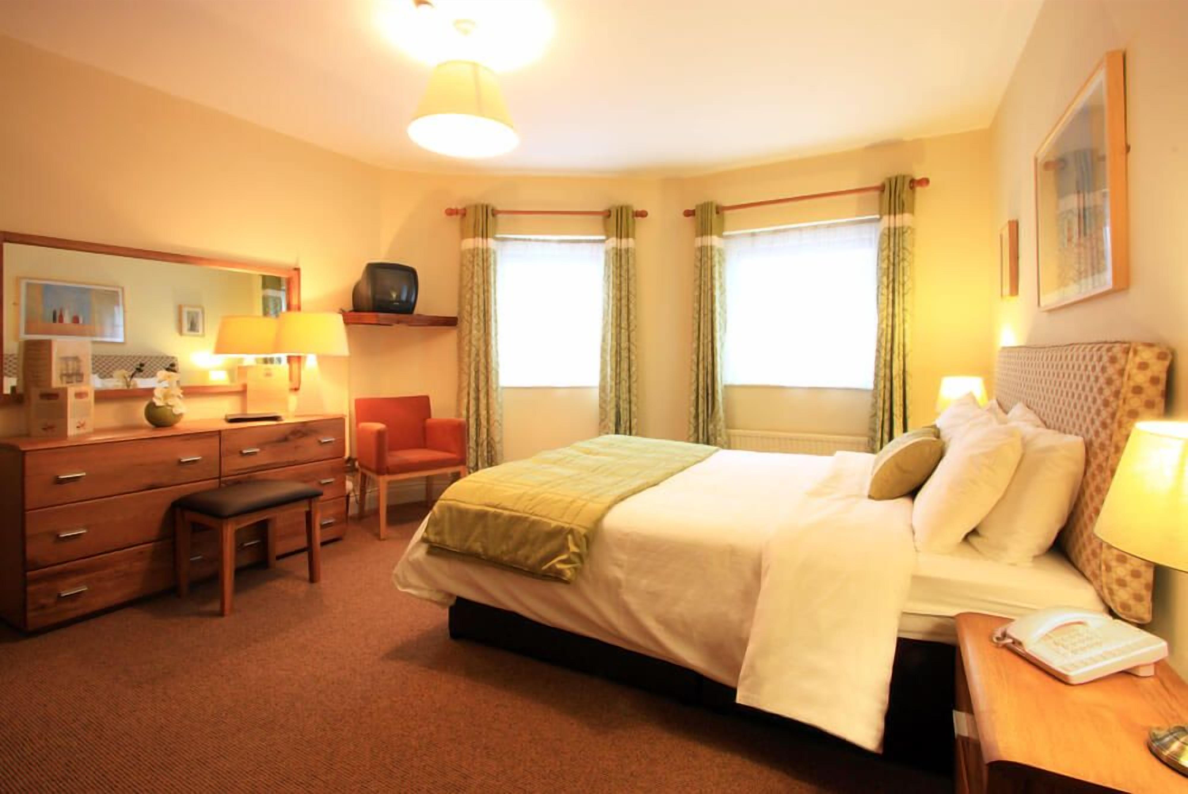 A hotel room with a bed, a desk, a chair, a dresser, a mirror, and two windows with curtains.