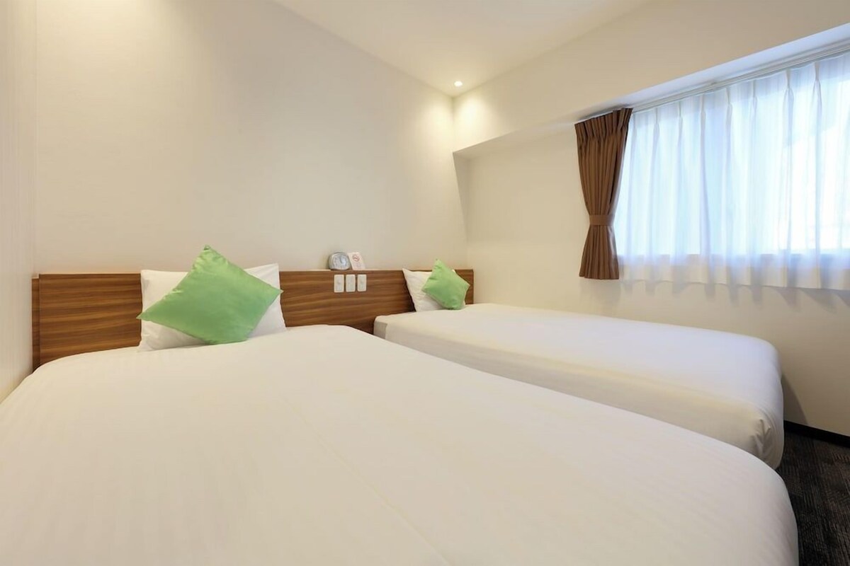A hotel room with two beds, a wooden headboard, a window with curtains, and a small table with a clock and a photo frame.