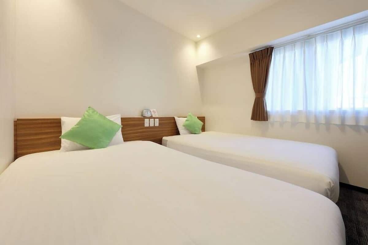 Habitació d'hotel amb dos llits, un panell de capçalera de fusta, una finestra amb cortines i una taula petita amb un rellotge i un marc de fotos.