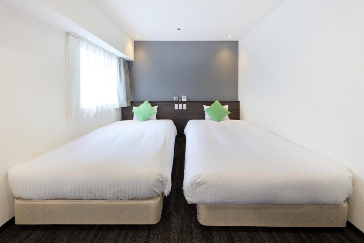 A hotel room with two single beds, a dark headboard, a window with curtains, and a grey wall.