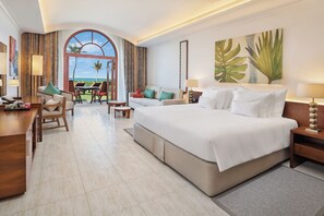 A hotel room with a large bed, a desk, a chair, a sofa, a dining table, and a view of the ocean.