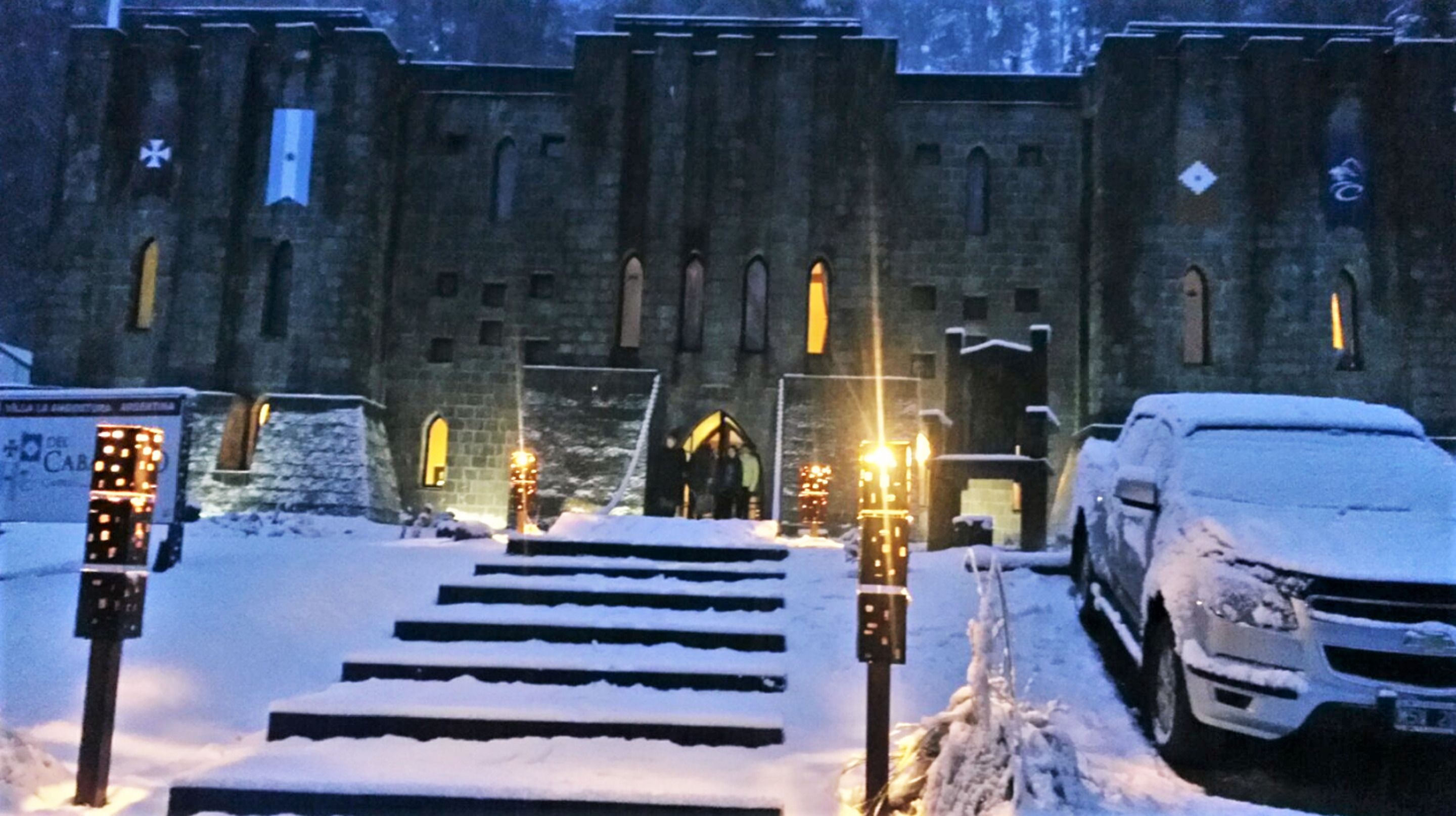 Un edificio cubierto de nieve con una escalera que lleva a una puerta, un SUV estacionado y farolas iluminadas.