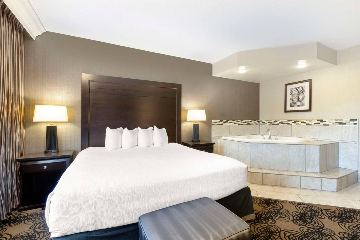 Quarto de hotel com uma cama grande, duas luminárias de cabeceira, banheiro com banheira e painel de azulejos.