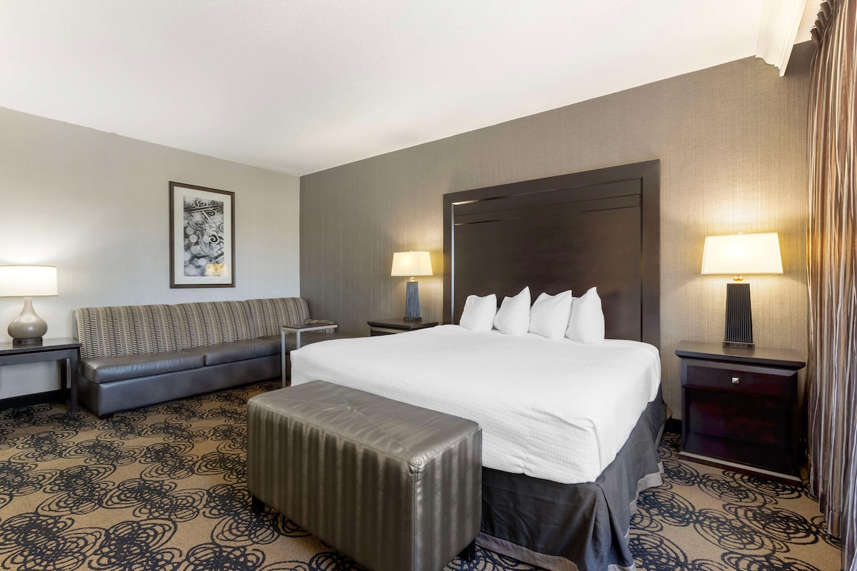 A hotel room with a large bed, a sofa, an ottoman, two nightstands, and a wall-mounted lamp.