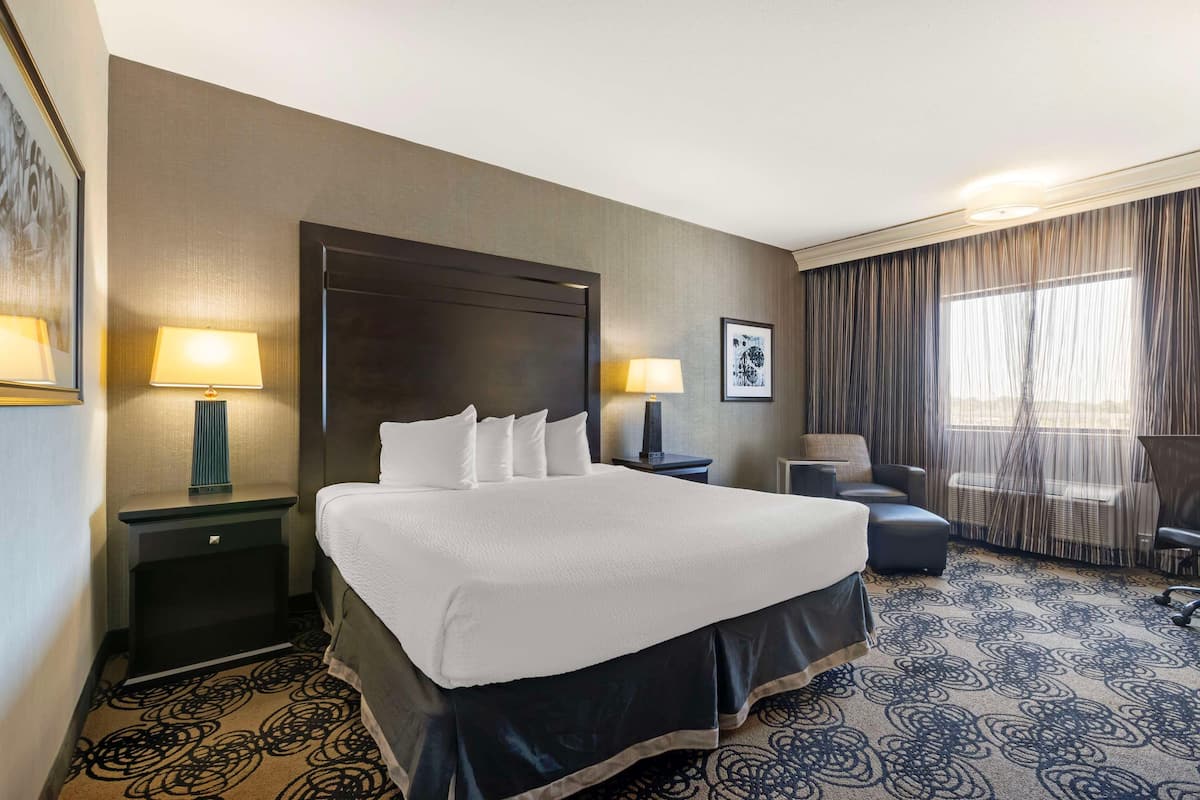 A hotel room with a large bed, two bedside tables with lamps, a chair, a desk, and a large window with curtains.