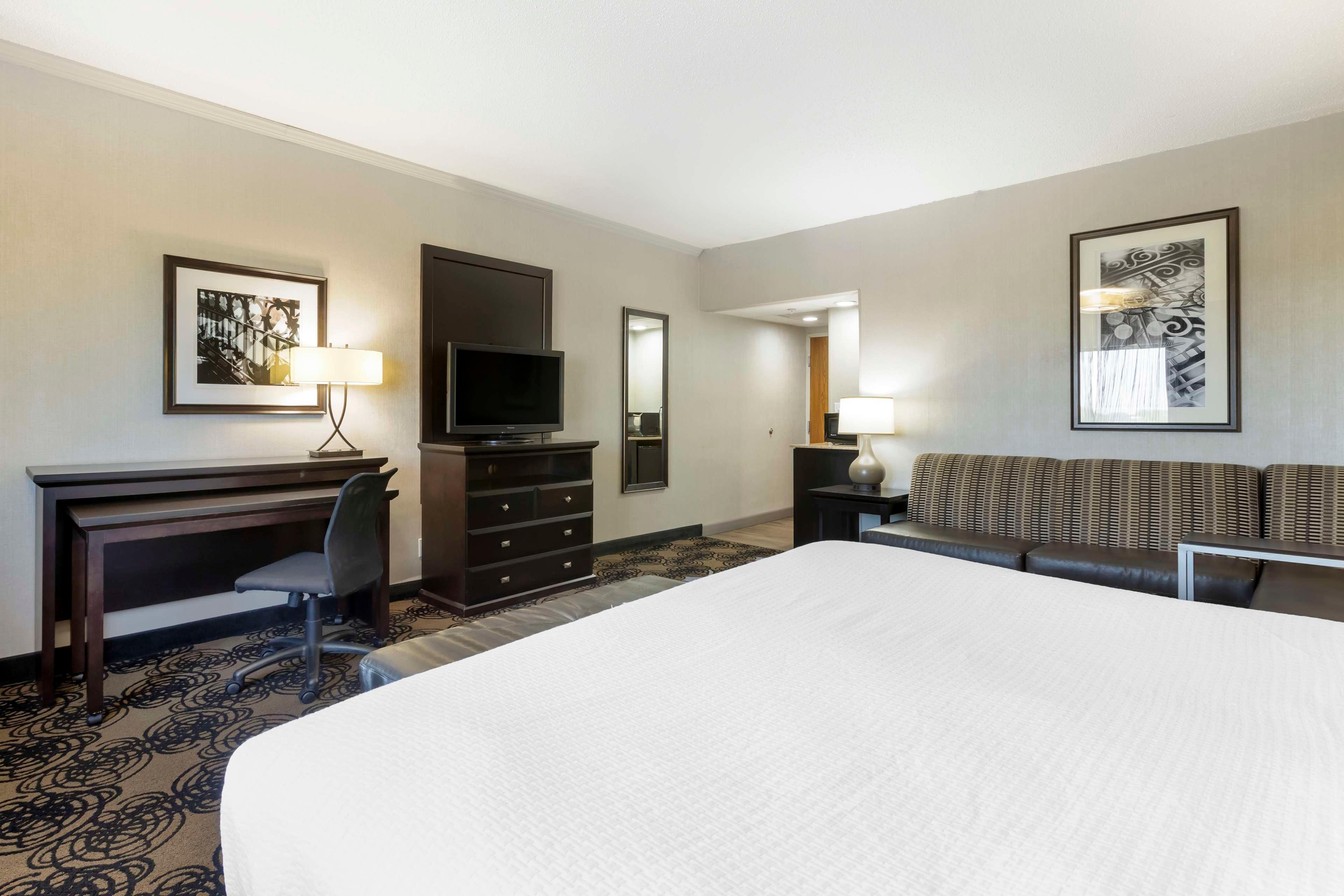 A hotel room with a large bed, a desk with a chair, a television, a sofa, and two framed artworks on the walls.