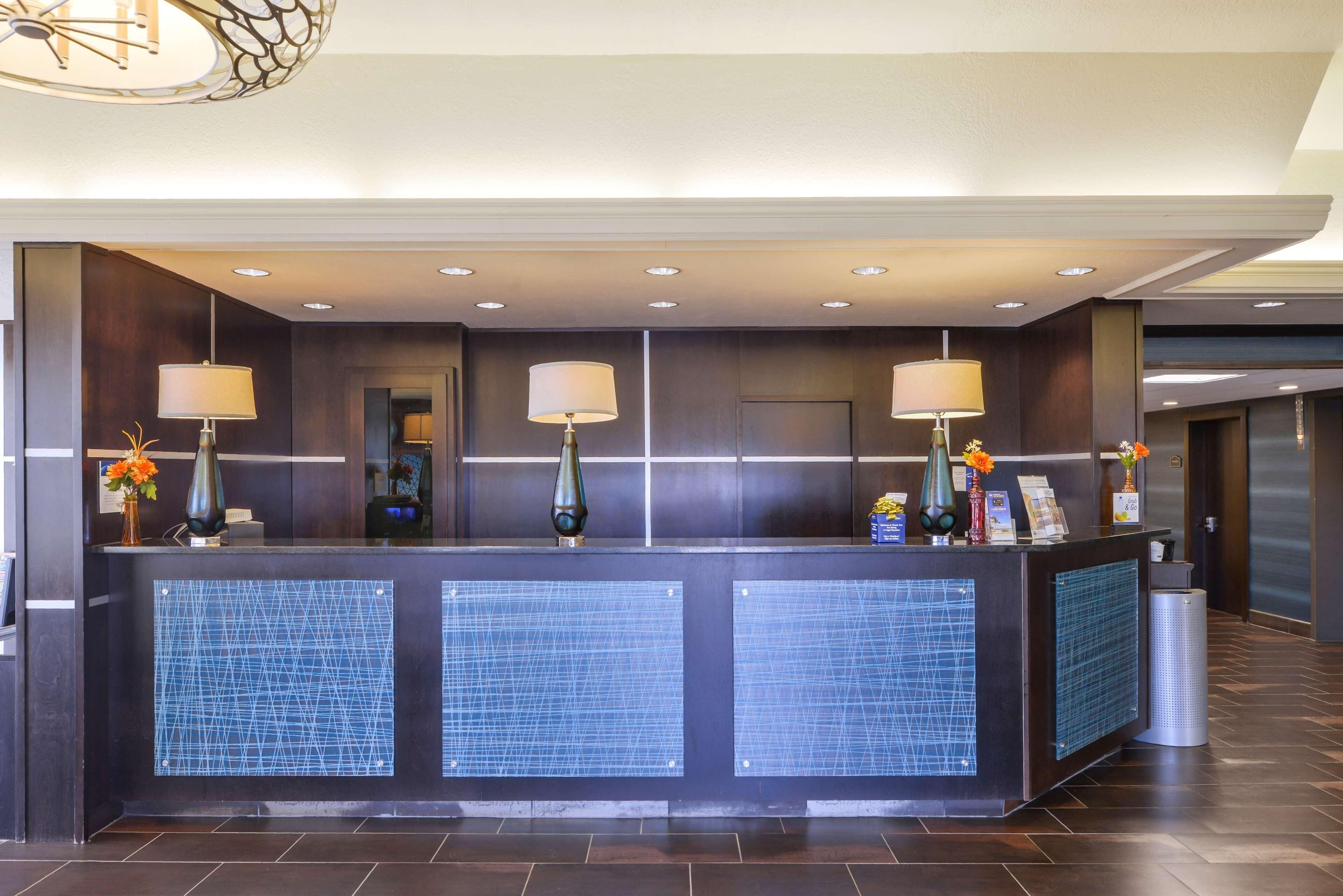 Uma receção de hotel com design moderno, destacando acabamento em madeira escura, uma vitrine em painel de vidro e três luminárias fixadas na parede.