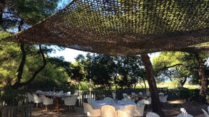 Un espace de restauration en plein air avec des tables rondes et des chaises blanches, à l’ombre d’un auvent en osier.