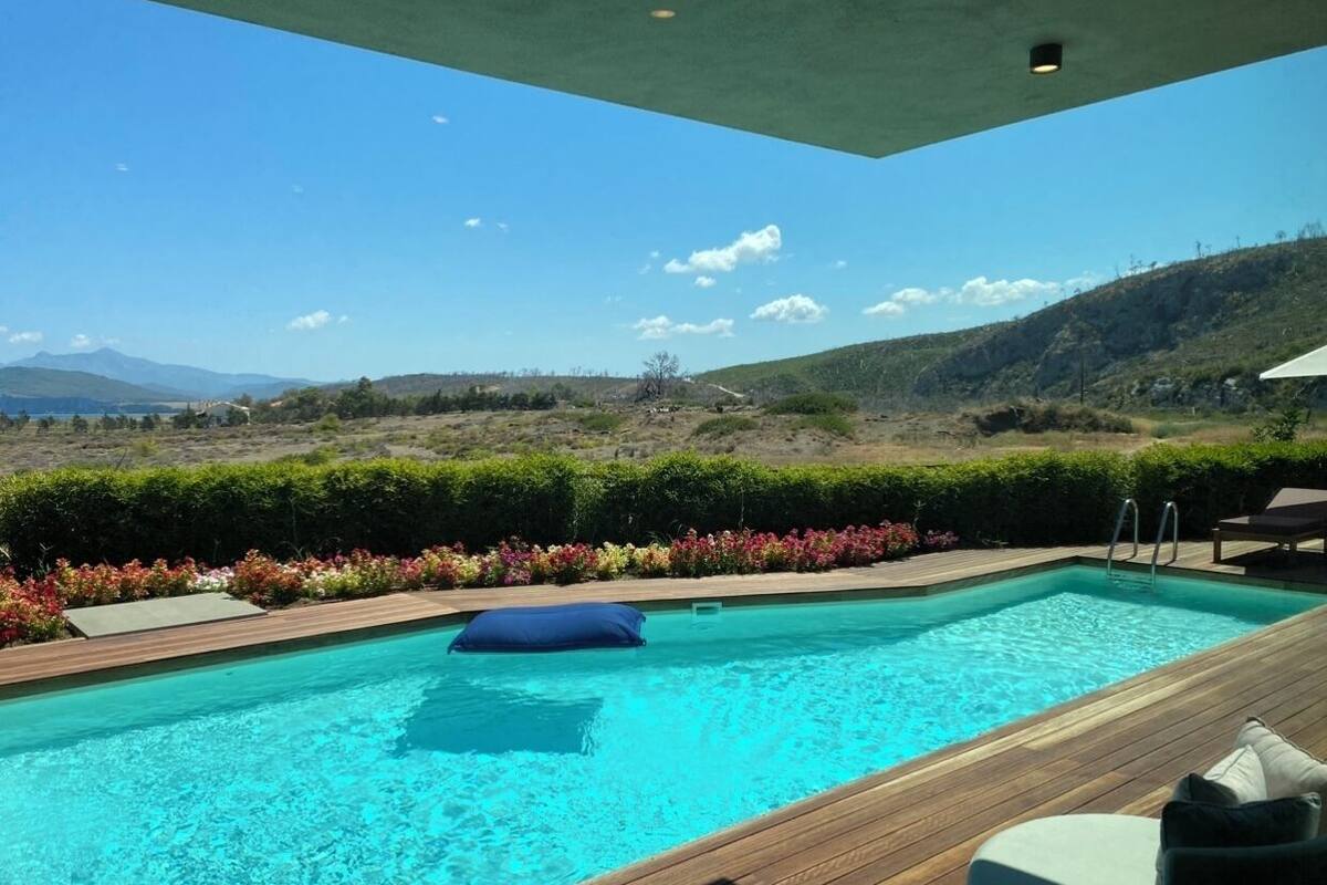 A rectangular swimming pool with a wooden deck, surrounded by colorful flowers and a view of hills and mountains.