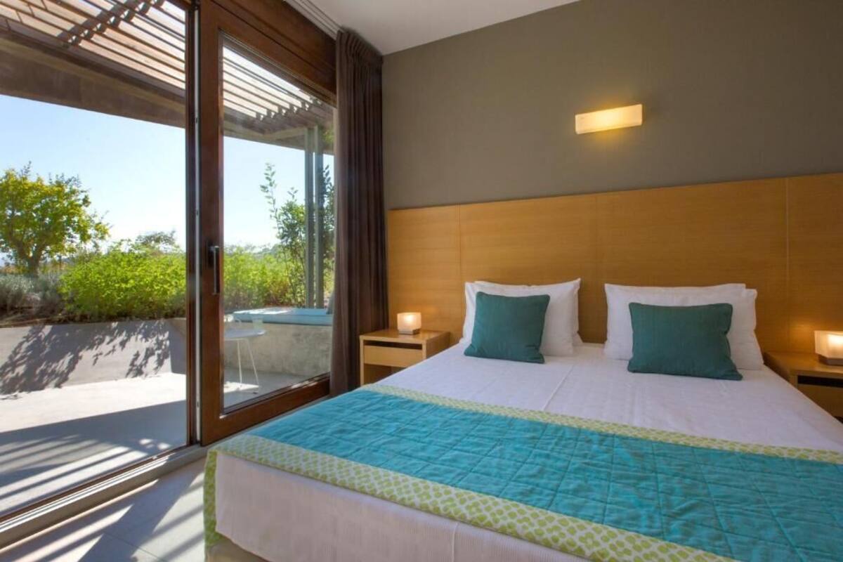 A bedroom with a large bed, two bedside tables, a nightstand, a window with curtains, and a view of greenery outside.