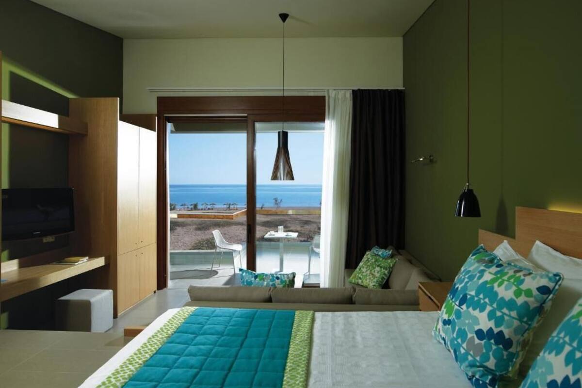 A hotel room with a bed, a TV, a sofa, a balcony with a view of the sea, and a dining area.