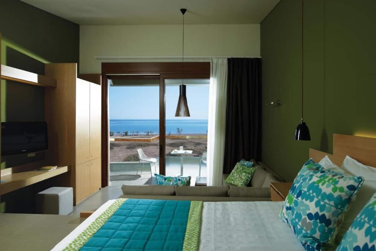 Une chambre d’hôtel comprenant un lit, une télévision, un canapé, un balcon avec vue sur la mer et un coin repas.