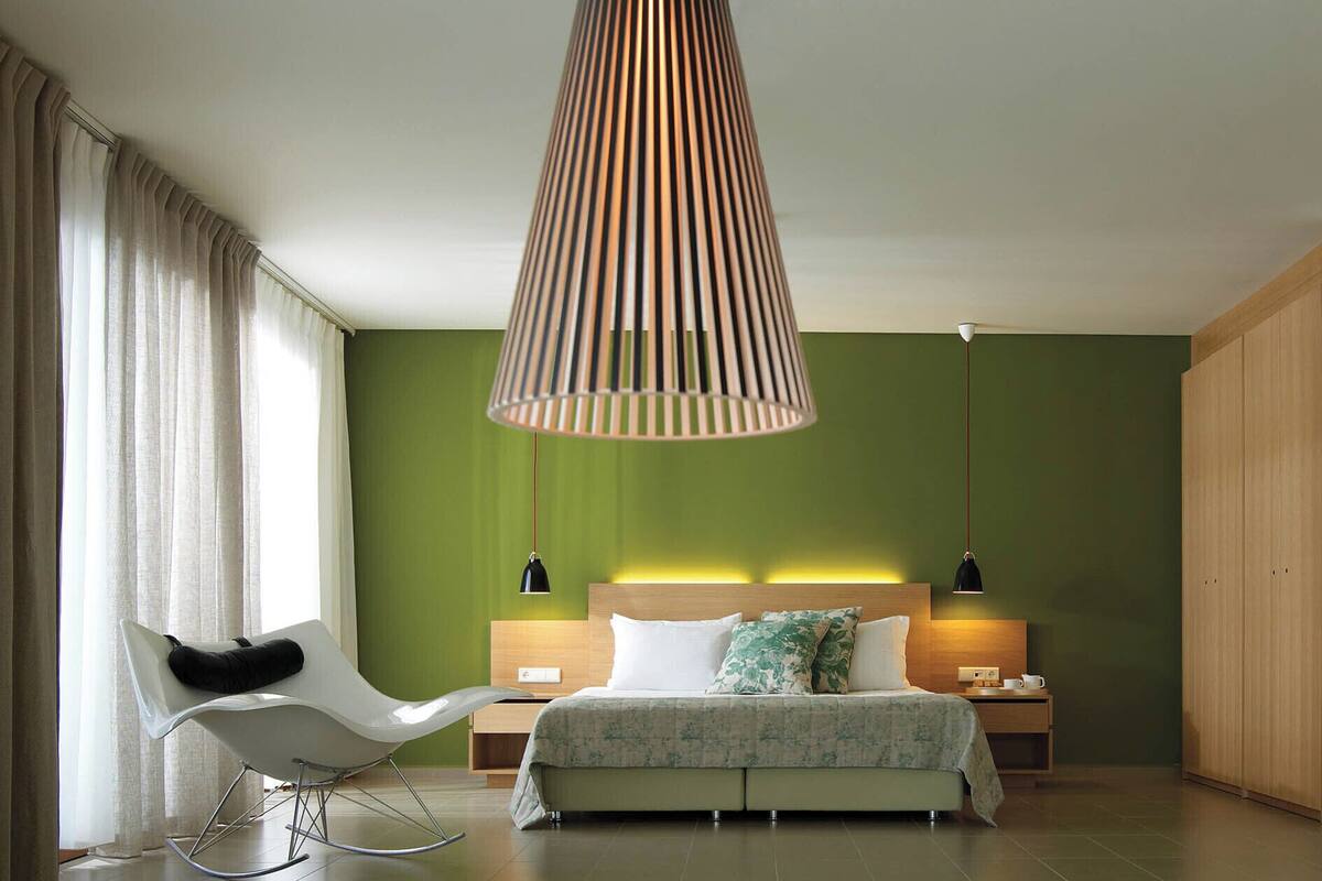 A modern bedroom with a green wall, a bed with white and green bedding, a white rocking chair, and a hanging lamp.