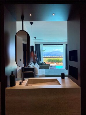 A modern hotel room with a large window, a bed, a bedside table, a mirror, and a view of a pool and mountains. - Thalatta Seaside Hotel (Mantoudi-Limni-Agia Anna)