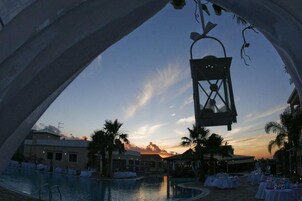 Zona de piscina con vista al atardecer, palmeras y un farol colgante.