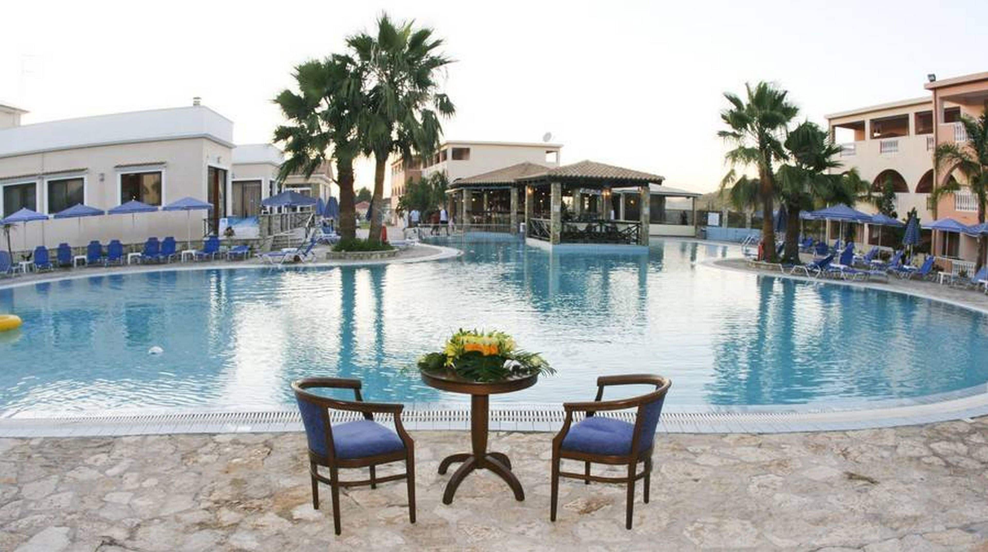 A swimming pool with lounge chairs and umbrellas, surrounded by palm trees and buildings.